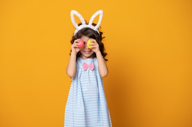 Fille enfant drôle portant des oreilles de lapin et tenant un oeuf de Pâques coloré devant ses yeux sur fond jaune.