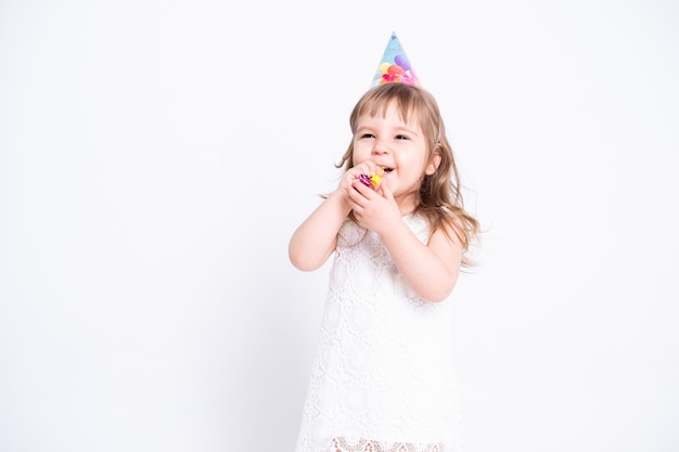 Fille enfant drôle en robe blanche et chapeau d'anniversaire soufflant dans le sifflet sur le mur blanc