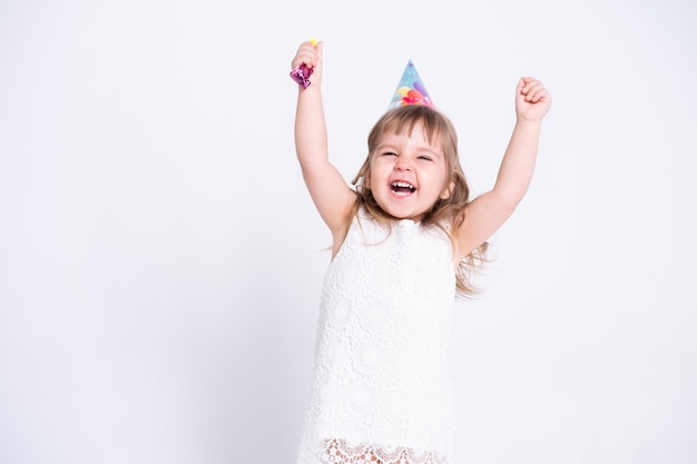 Fille enfant drôle en robe blanche et chapeau d'anniversaire soufflant dans le sifflet sur le mur blanc