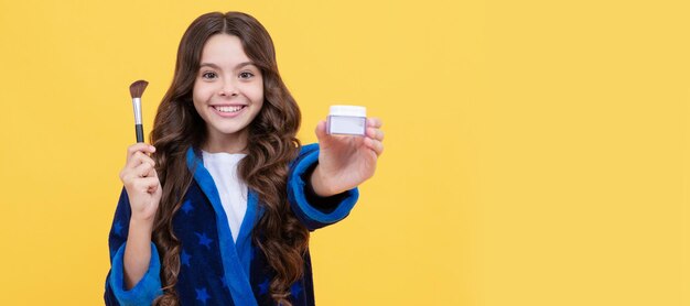 Fille enfant heureuse en peignoir confortable se toilettant avec un pinceau à poudre présentant le produit Cosmétiques adolescent