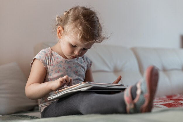 Une fille d'enfants en vêtements de maison est assise sur le canapé et apprend sur une tablette. Formation à distance et gadgets