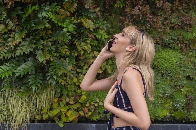 Photo fille d'entrepreneur dans un parc d'affaires sur un mur vert de plantes, jeune blonde souriante passant un appel avec le téléphone