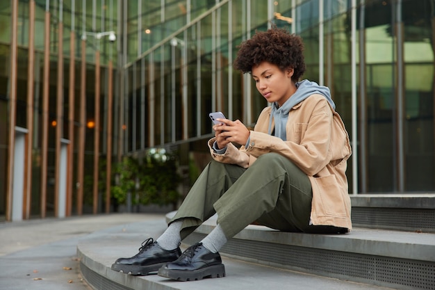 une fille envoie un message sms à un ami sur un smartphone porte des vêtements élégants est assise sur des marches près d'un bâtiment en verre moderne recherche des informations