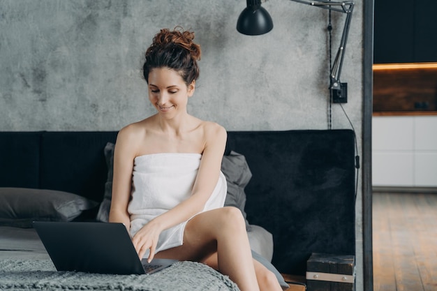 Une fille espagnole indépendante enveloppée dans une serviette après le bain est assise devant un ordinateur portable sur son lit et souriante