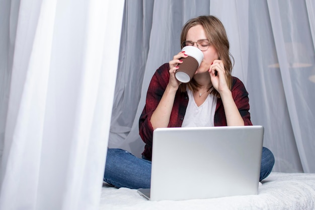 Une fille est allongée dans son lit avec un ordinateur portable et boit un café le matin une pigiste travaille à la maison