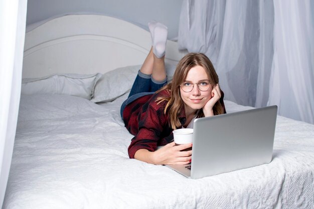 Une fille est allongée dans son lit avec un ordinateur portable et regarde une vidéo avec du café. Une pigiste travaille à la maison