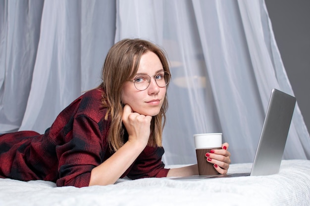 Une fille est allongée dans son lit avec un ordinateur portable et regarde une vidéo avec une pigiste de café