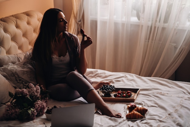 Une fille est assise au lit le soir en regardant un ordinateur portable et en mangeant des fraises, une fille au lit mange des bonbons avant d'aller se coucher