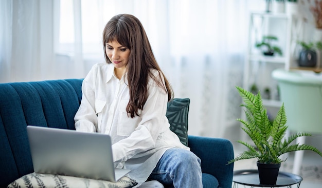 La fille est assise sur le canapé et travaille sur un ordinateur portable dans l'appartement