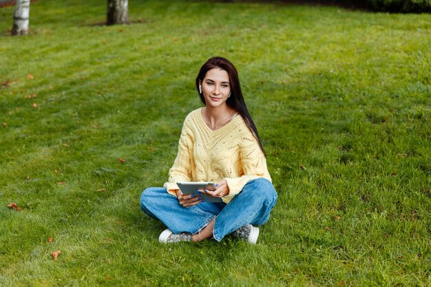 La fille est assise dans le parc et est engagée avec l'iPad. jeune brune est assise sur l'herbe et se penche sur la tablette. fille dans le parc en automne.