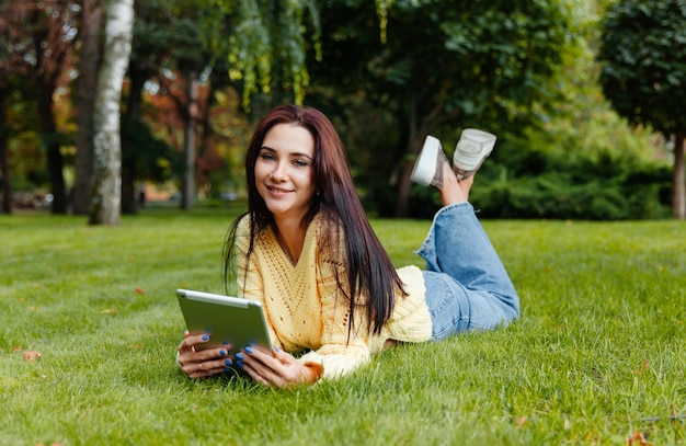 La fille est assise dans le parc et est engagée avec l'iPad. jeune brune est assise sur l'herbe et se penche sur la tablette. fille dans le parc en automne.