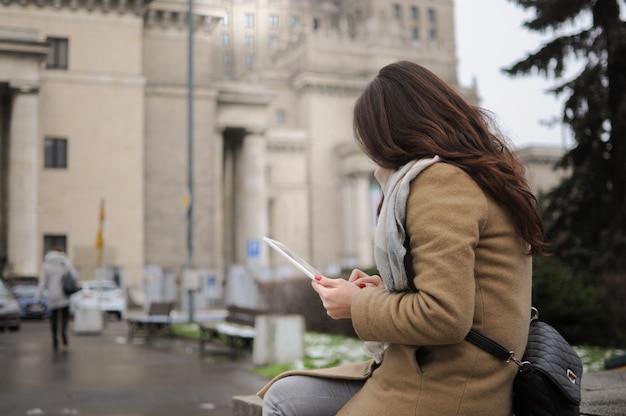 La fille est assise et envoie des SMS dans sa tablette à l'extérieur en hiver. SMS en ligne.