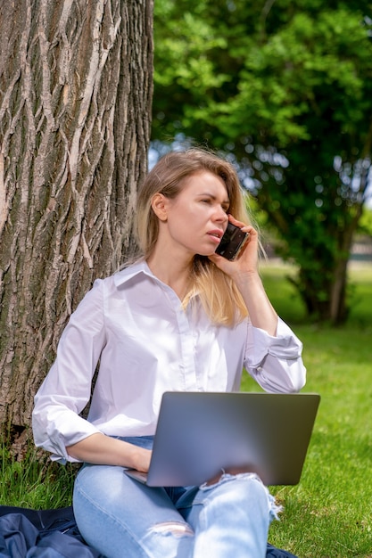 Photo une fille est assise sur l'herbe près d'un arbre dans un parc de pays avec ordinateur portable