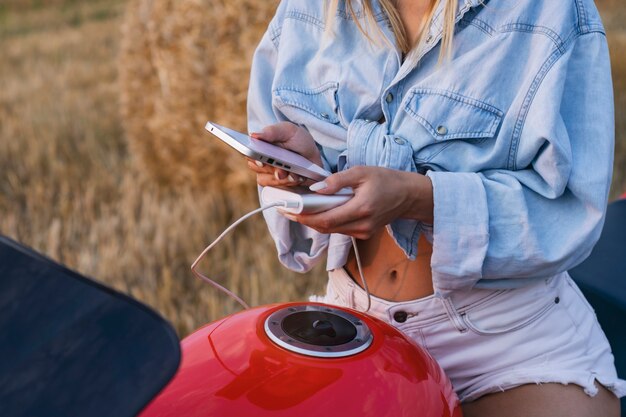 Une fille est assise sur une moto et tient une maquette d'un smartphone avec un écran blanc. Power Bank charge votre téléphone dans le contexte de la nature.