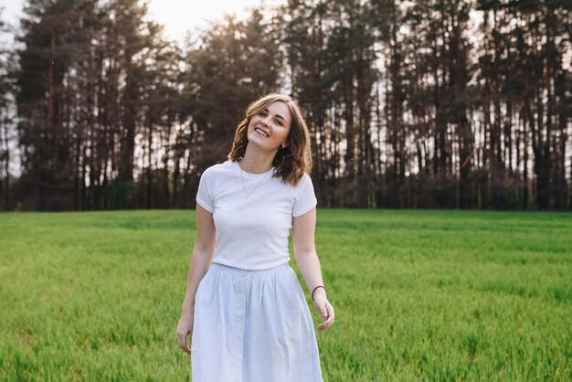 La fille est blonde, cheveux bruns, dans une chemise blanche et une jupe midi bleue. Marcher dans le champ, à travers l'herbe verte. Portrait d'une fille. Positif et souriant.