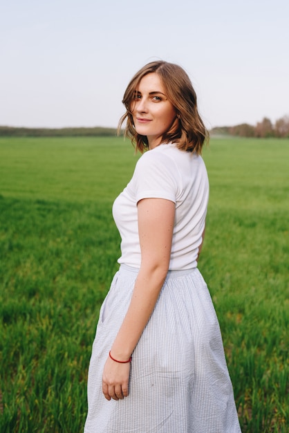 La fille est blonde, cheveux bruns, dans une chemise blanche et une jupe midi bleue. Marcher dans le champ, à travers l'herbe verte. Portrait d'une fille. Positif et souriant.