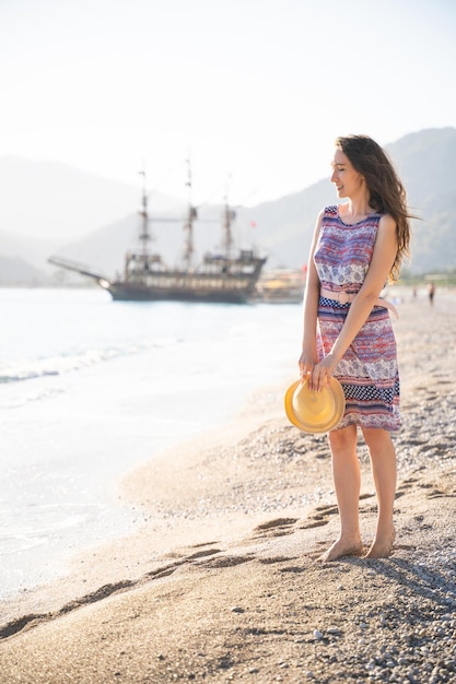 Fille en été en robe et chapeau sur le rivage de l'océan et grand navire amarré