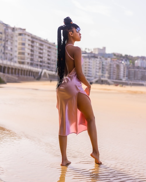 Fille ethnique dominicaine avec des tresses avec une belle robe rose. Posant à la mode profitant de l'été sur la plage au coucher du soleil