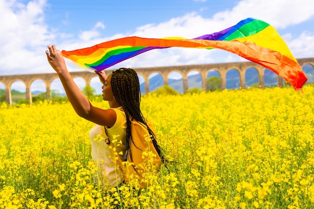 Une fille ethnique noire avec des tresses tenant le drapeau LGBT dans un champ de fleurs jaunes