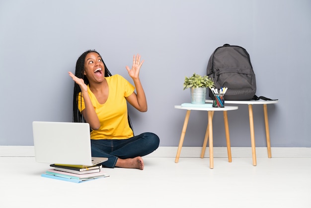 Fille étudiante adolescente afro-américaine avec de longs cheveux tressés, assise sur le sol avec une expression faciale surprise