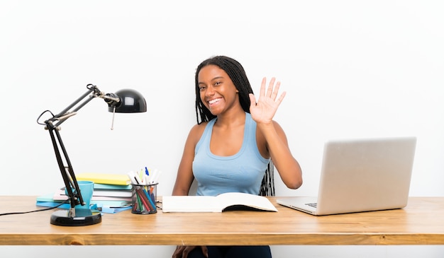 Fille étudiante adolescente afro-américaine avec de longs cheveux tressés sur son lieu de travail, saluant avec la main avec une expression heureuse