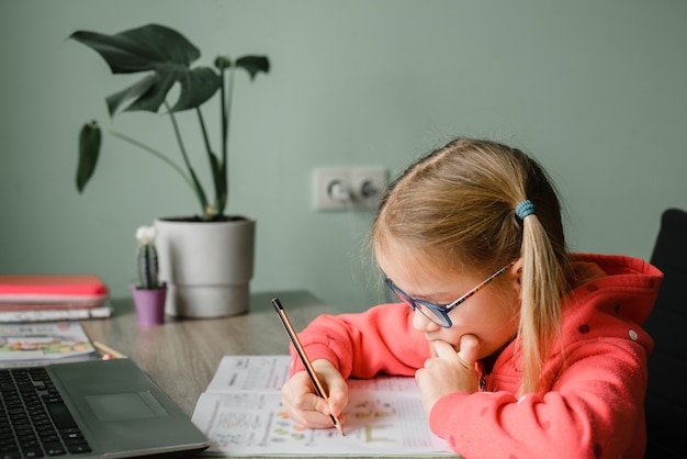 Fille étudiante junior dans les yeux écrivant dans son cahier tout en étudiant en ligne à la maison, étude du concept de la maison