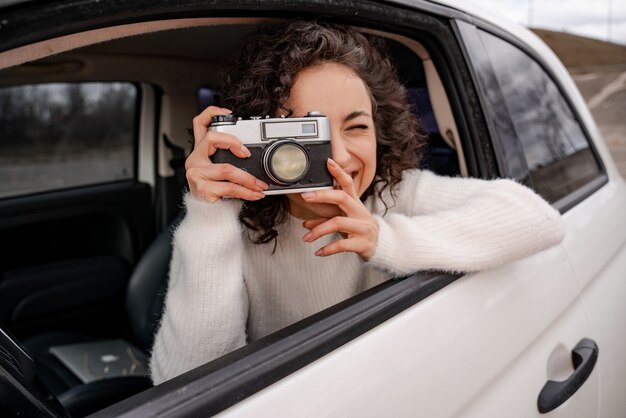 Une fille européenne prend une photo sur un vieil appareil photo à partir d'une automobile personnelle. La jeune belle femme bouclée est concentrée et souriante