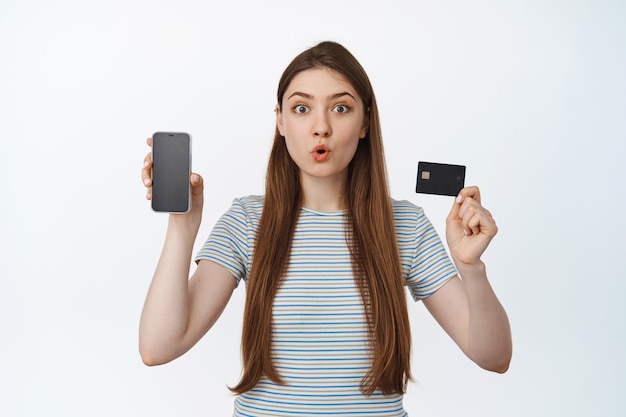 Une fille excitée et surprise montre une carte de réduction de crédit et un écran de téléphone portable, une interface d'application pour smartphone, debout sur blanc.