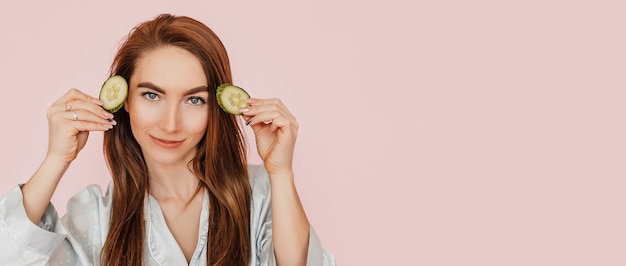 Une fille fabrique des masques de beauté pour le visage faits maison. Concombres pour la fraîcheur de la peau autour des yeux. La femme prend soin d'une peau jeune. Modèle riant et s'amusant au spa sur fond rose