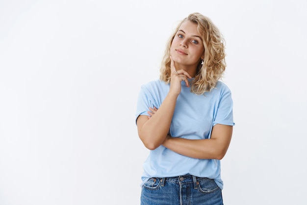 Fille faisant le choix dans la boutique debout dans une pose hmm réfléchie avec le doigt sur la mâchoire inclinant la tête et souriant en regardant la caméra choisir le produit en pensant et en prenant une décision sur le mur blanc