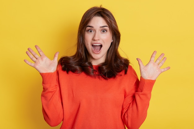 Fille faisant un geste de surprise, criant quelque chose et agitant joyeusement les paumes levées, posant joyeusement avec des expressions enthousiastes contre le jaune, portant un pull orange.