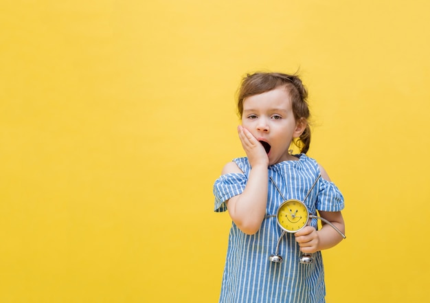 Fille fatiguée sur un espace jaune est titulaire d'un réveil. Une petite fille bâille sur un espace jaune. Jolie fille avec des nattes dans un chemisier rayé.