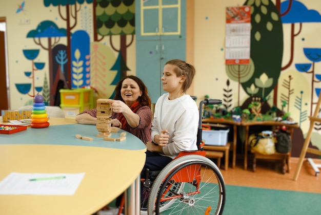 Une fille en fauteuil roulant joue à des jeux de société assise à une table Une psychologue est engagée avec une personne handicapée