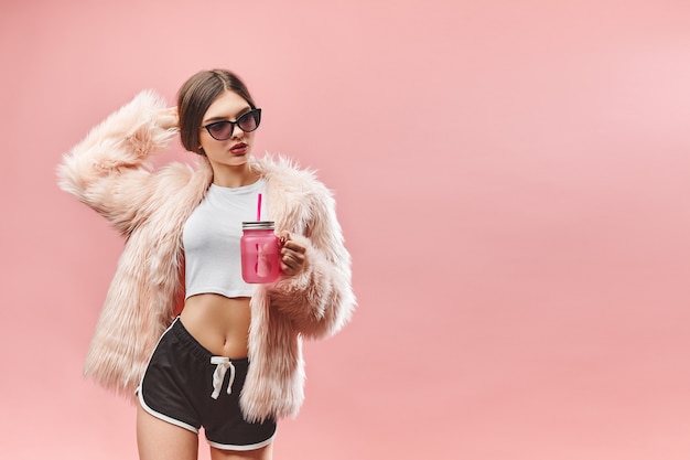 Fille en faux manteau de fourrure rose short noir haut blanc et lunettes de soleil tient une boisson rose avec les yeux fermés...