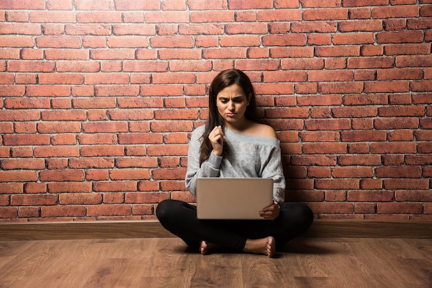 Fille ou femme indienne assise avec un ordinateur portable ou utilisant un ordinateur portable sur un plancher en bois contre un mur de briques rouges