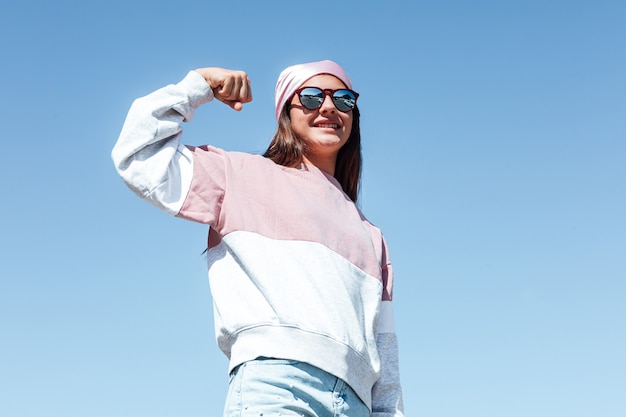 Fille femme avec des lunettes de soleil et un foulard rose sur la tête, serre son bras en signe de force. Journée internationale du cancer du sein, avec le ciel comme arrière-plan.