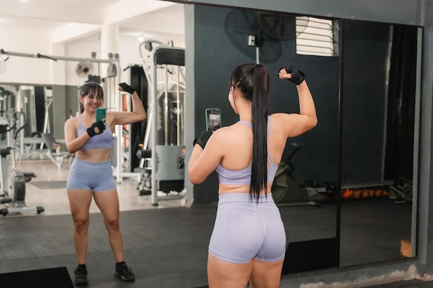 Une fille de fitness montrant des muscles en prenant des photos avec son smartphone devant le miroir du gymnase