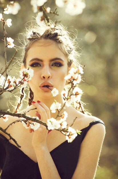 Fille et fleurs jolie femme mangeant des fleurs de printemps de cerise blanche ou d'abricot en fleurs a un maquillage à la mode sur le visage et des cheveux élégants ensoleillés en plein air sur fond naturel