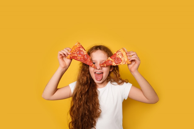 Une fille folle bouclée tient deux tranches de pizza près de ses yeux et lui tire la langue.