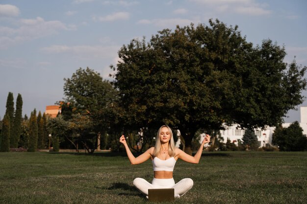 Une fille en forme est assise en position du lotus et médite en ligne avec un ordinateur portable Méditation et relaxation à l'extérieur