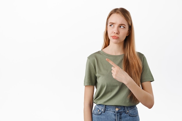 Fille froncée réfléchie pointant du doigt, regardant vers la gauche avec des doutes, regardant incertaine l'espace de copie, debout sur un mur blanc