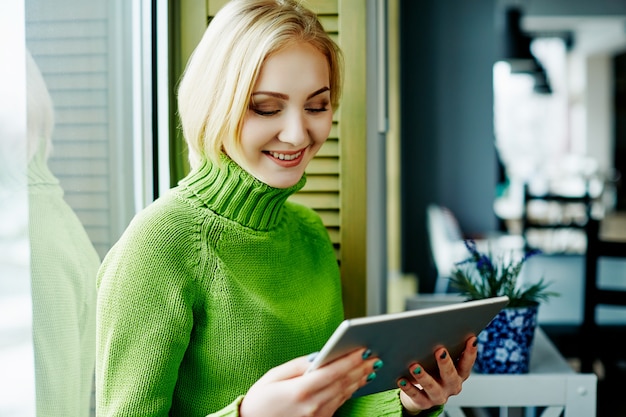 Fille gaie aux cheveux clairs portant un pull vert assis dans un café avec tablette, concept indépendant, achats en ligne, portrait.