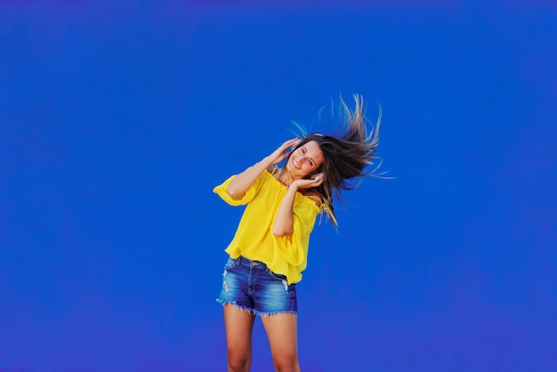 Fille gaie en chemise jaune debout contre le mur bleu avec un casque sur la tête.