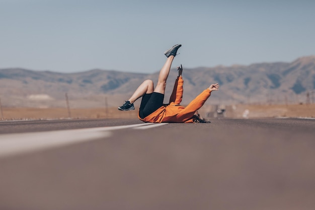 Une fille gaie dans un chandail à capuchon orange pose de façon brillante allongée sur la route sur fond de grandes hautes montagnes rocheuses