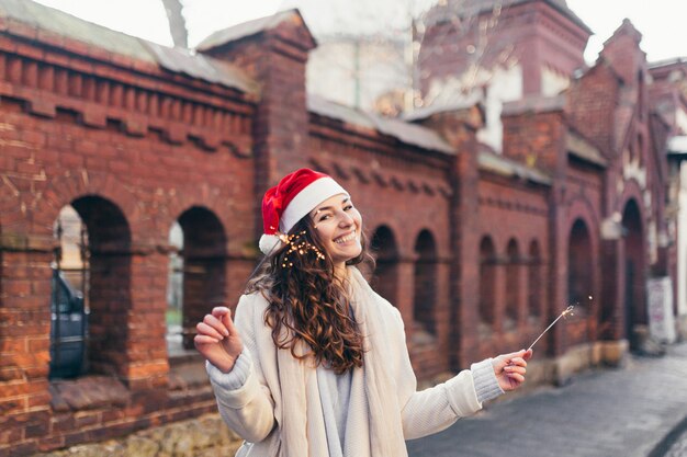 Fille gaie dans un chapeau de Noël souriant un dans la ville tenant des cierges magiques