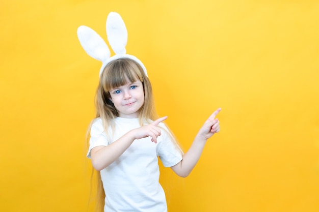 Fille gaie avec des oreilles de lapin sur la tête sur un fond jaune Drôle enfant heureux pointe du doigt un espace vide copie espace pour la maquette de texte