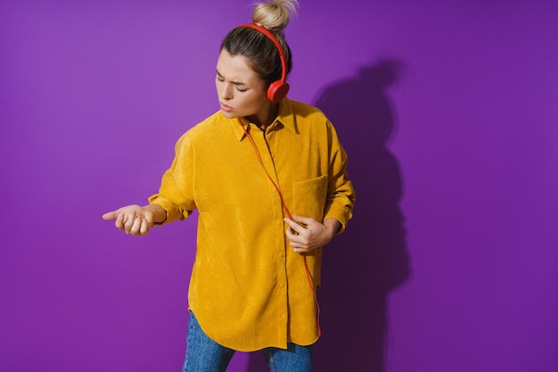 Fille gaie portant une chemise jaune écoutant de la musique à l'aide d'un casque rouge et jouant de la guitare à air sur fond violet