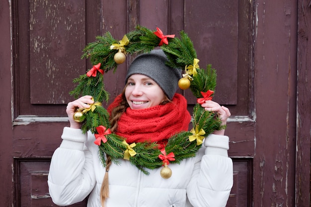 Fille en gants rouges et veste blanche tenant une couronne de Noël devant la porte