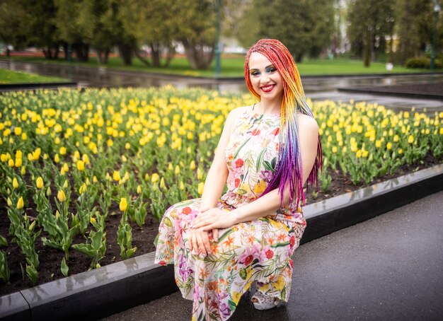 Fille géniale avec des tresses arc-en-ciel et un maquillage expressif à paillettes. Elle est assise et profite du temps chaud du printemps dans un parc verdoyant avec de grands parterres de tulipes.