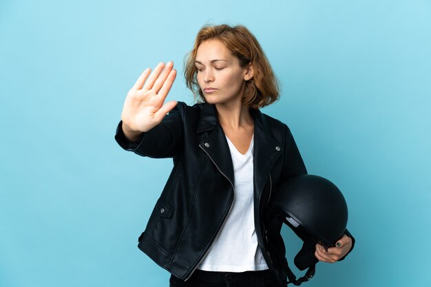 Fille géorgienne tenant un casque de moto isolé sur mur bleu faisant le geste d'arrêt et déçu